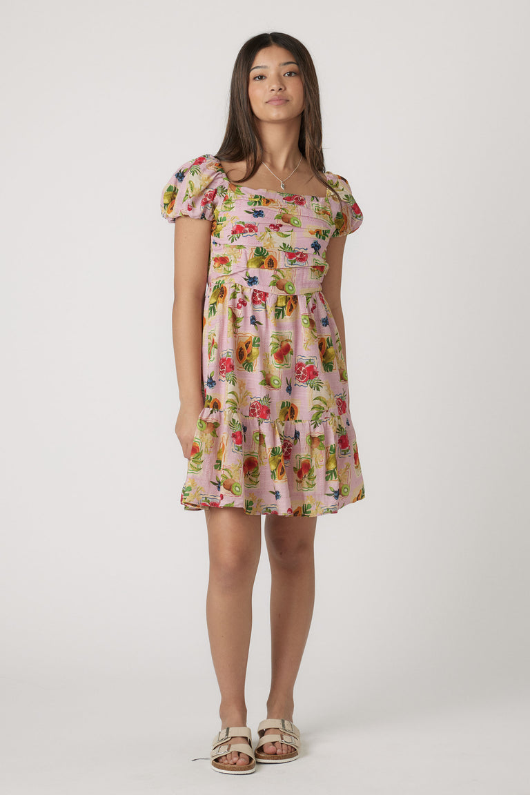 Girl wearing a pink mini dress with puff sleeves and colorful fruit print paired with cream sandals in a simple studio setting