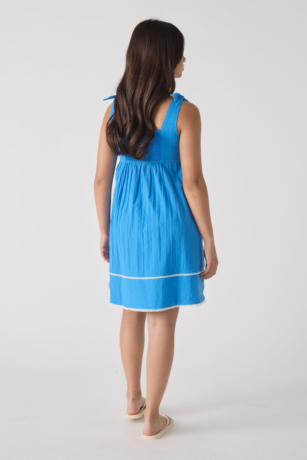 Young girl wearing a vibrant blue sleeveless mini dress with tie straps and delicate white trim, shown from behind.