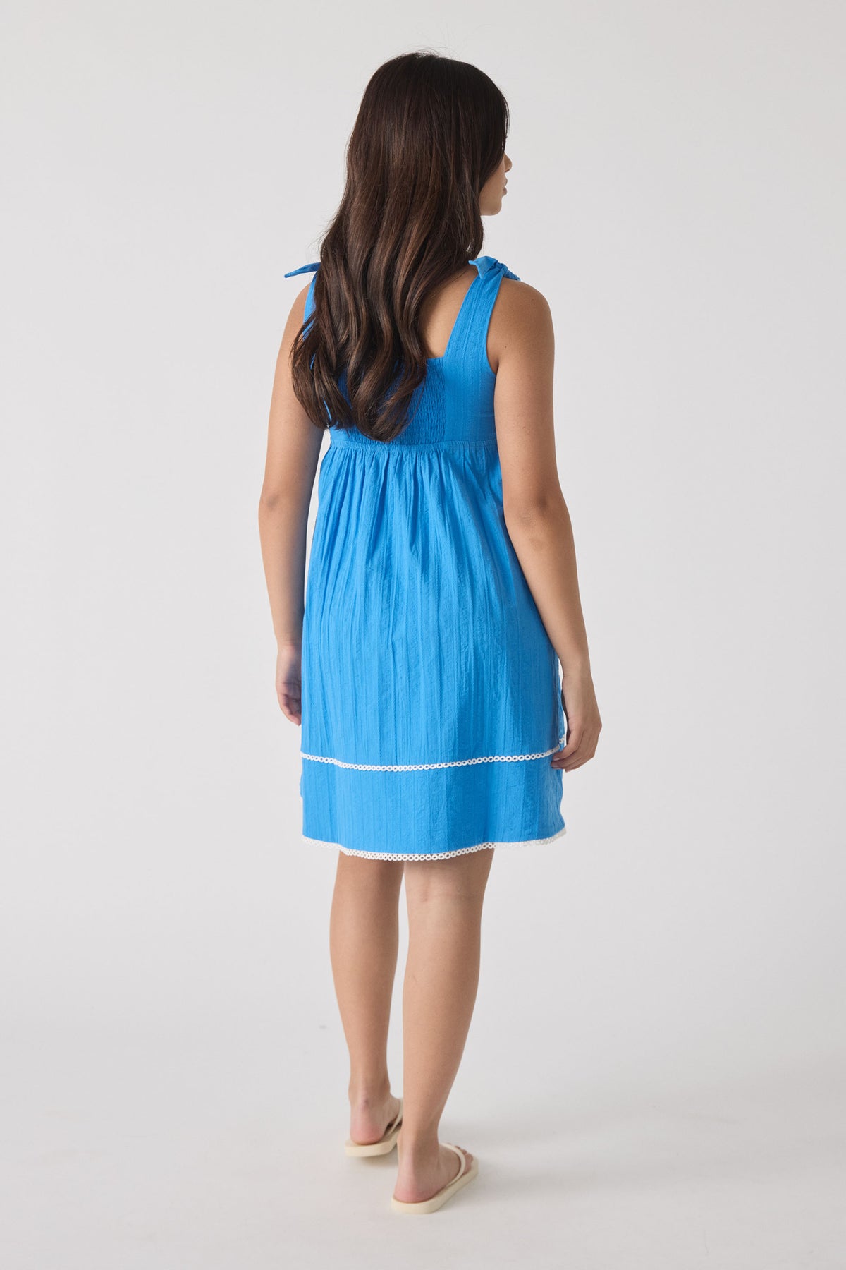 Young girl wearing a vibrant blue sleeveless mini dress with tie straps and delicate white trim, shown from behind.