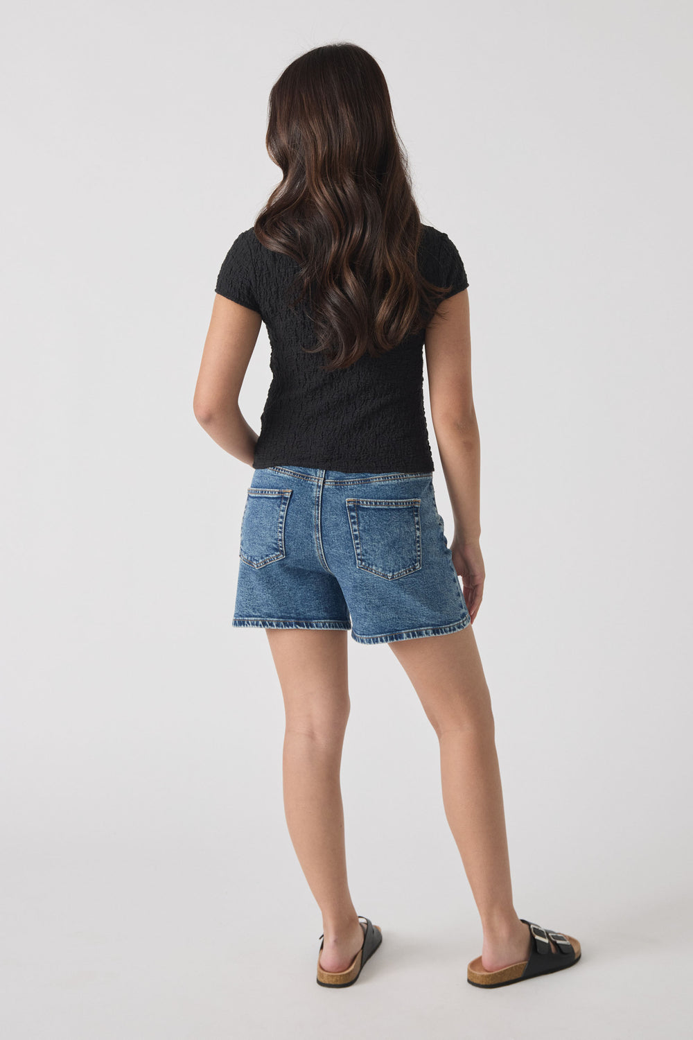Girl wearing mid-blue denim shorts with a straight hem and a fitted black textured short-sleeve top, shown from behind.