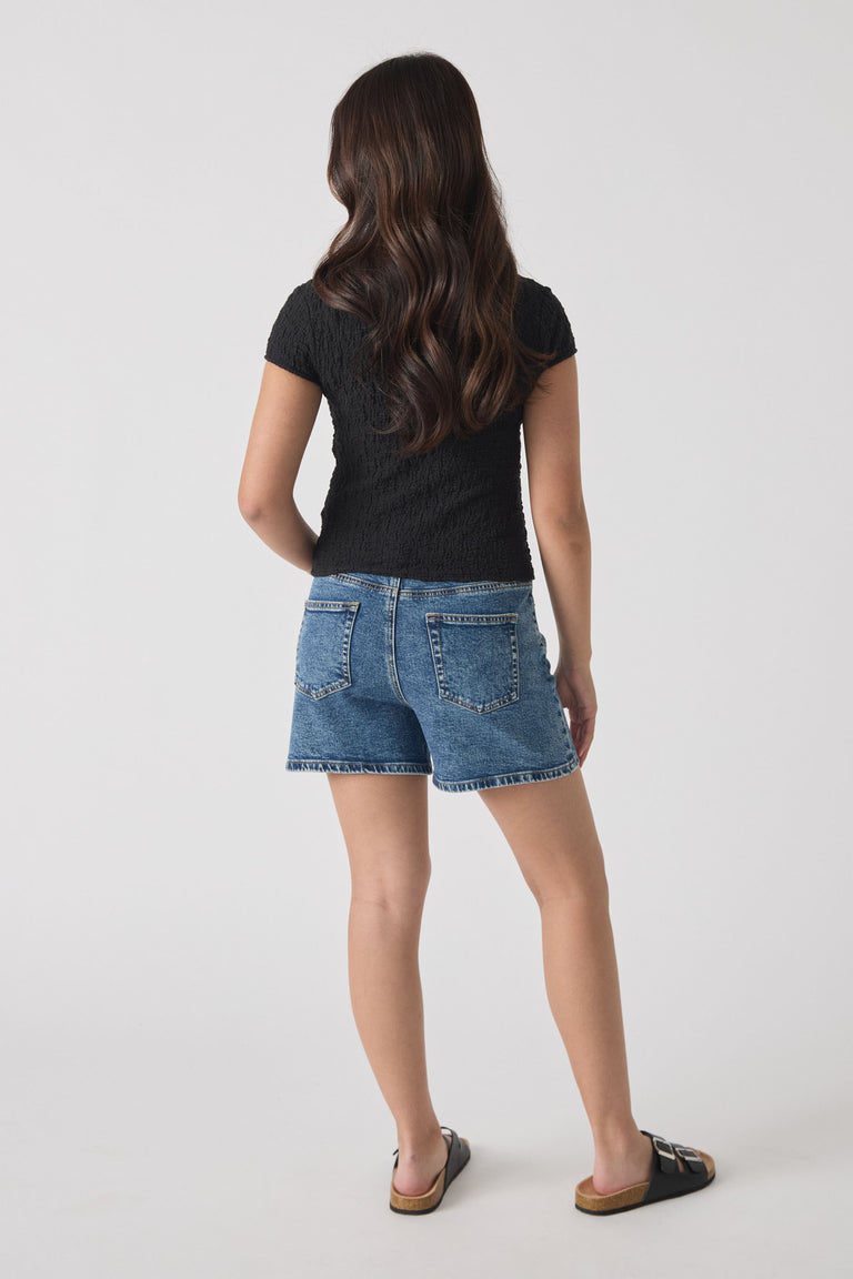 Girl wearing mid-blue denim shorts with a straight hem and a fitted black textured short-sleeve top, shown from behind.
