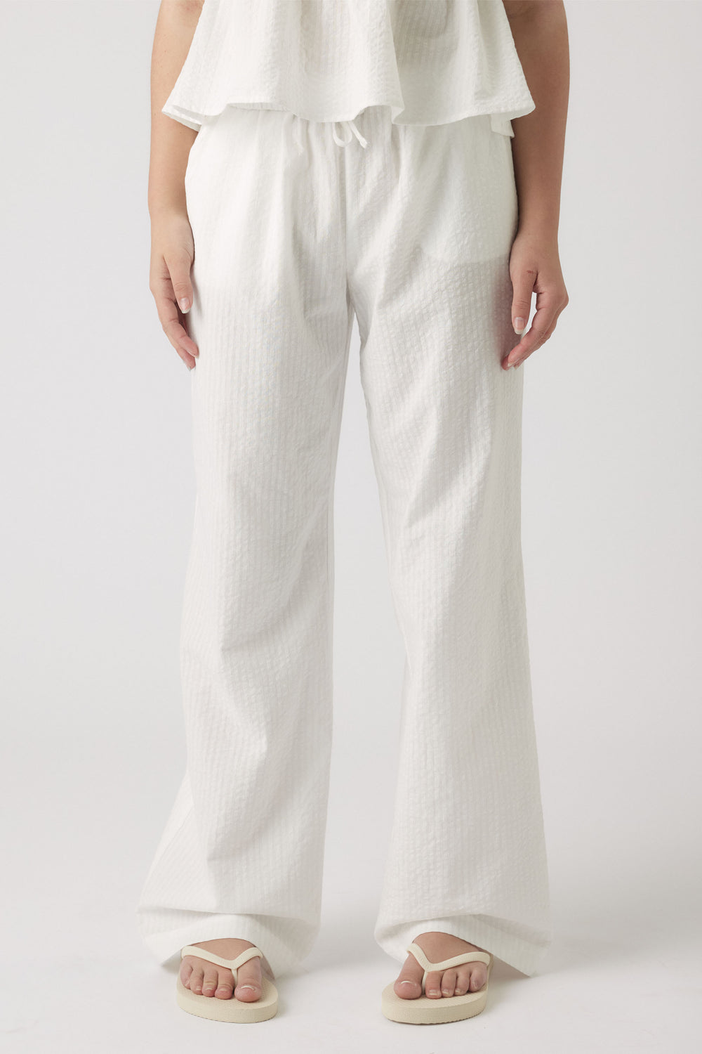Young girl wearing relaxed white textured summer pants with a drawstring waist paired with casual flip flops.