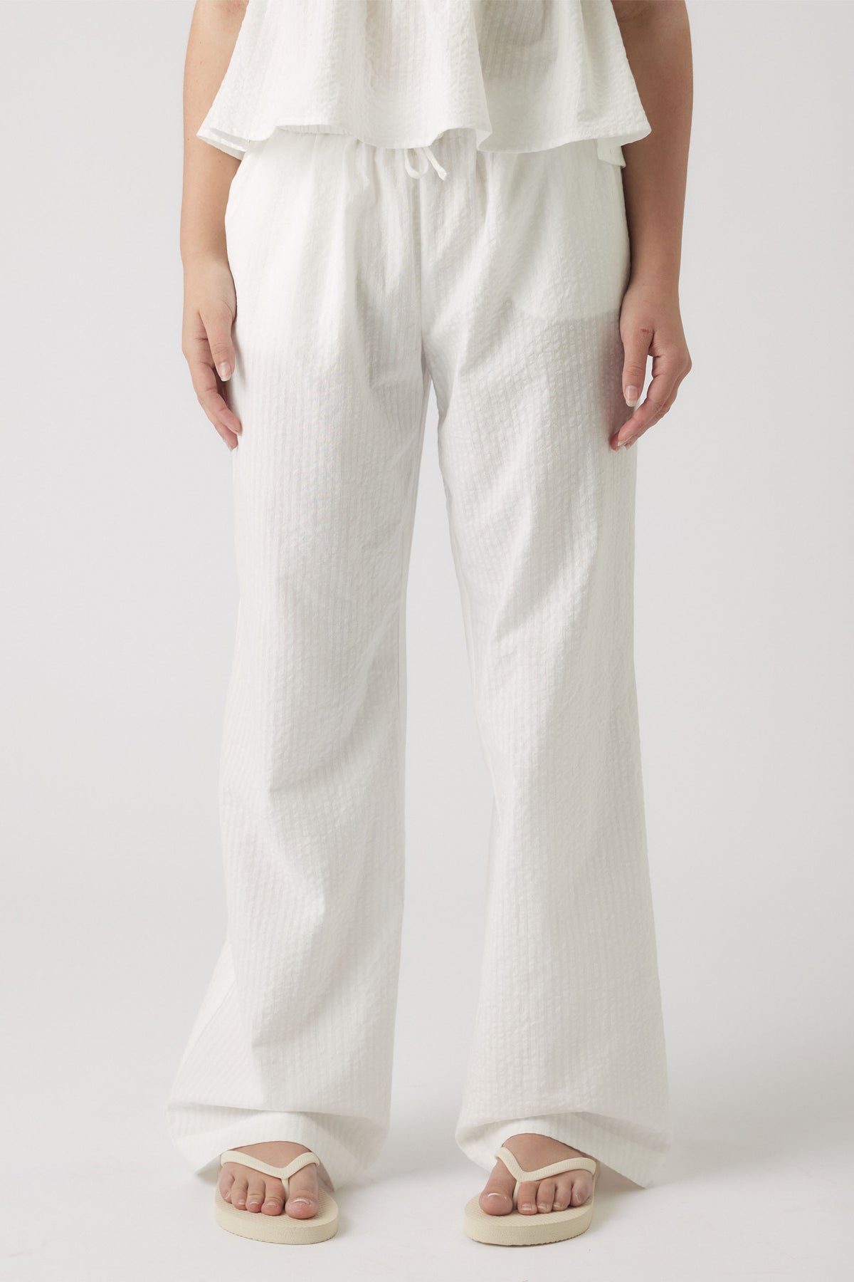 Young girl wearing relaxed white textured summer pants with a drawstring waist paired with casual flip flops.