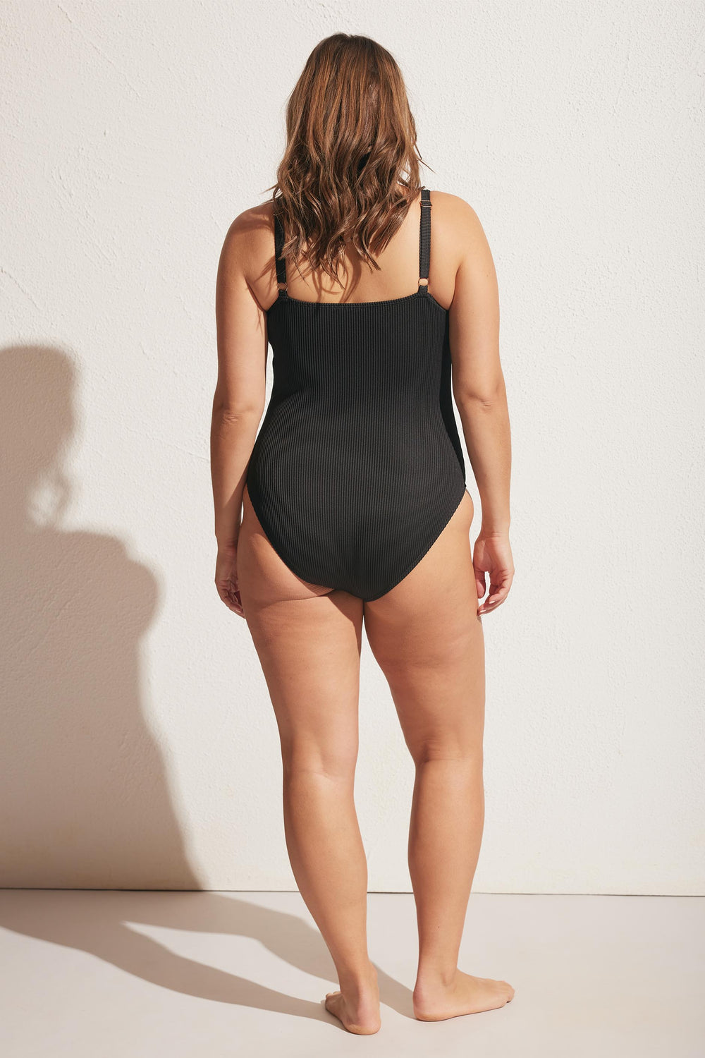 Woman wearing a black ribbed one-piece swimsuit with adjustable straps, shown from the back against a plain wall.