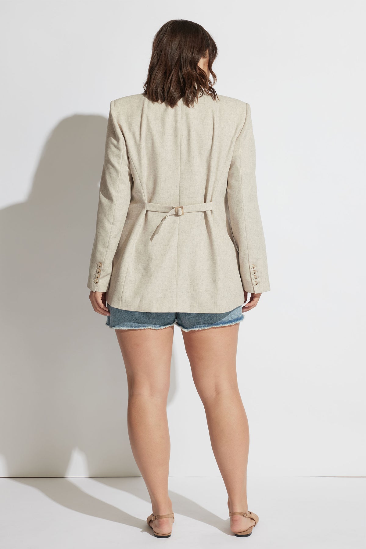 Woman wearing a beige tailored blazer with a cinched waist belt paired with blue distressed denim shorts and sandals