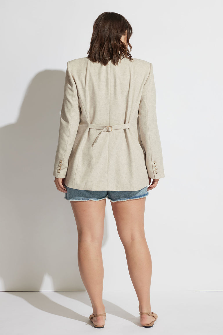 Woman wearing a beige tailored blazer with a cinched waist belt paired with blue distressed denim shorts and sandals