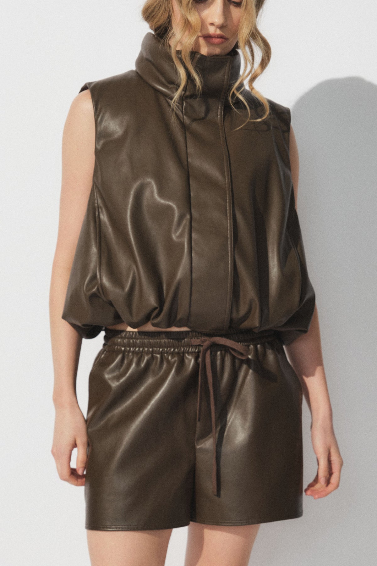 Woman wearing a sleeveless faux leather padded vest with matching brown faux leather shorts in a relaxed fit
