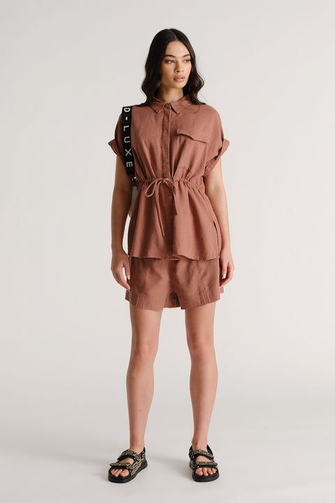 Woman wearing rust-colored linen blend shorts with a matching belted shirt and black strap sandals in a casual pose.