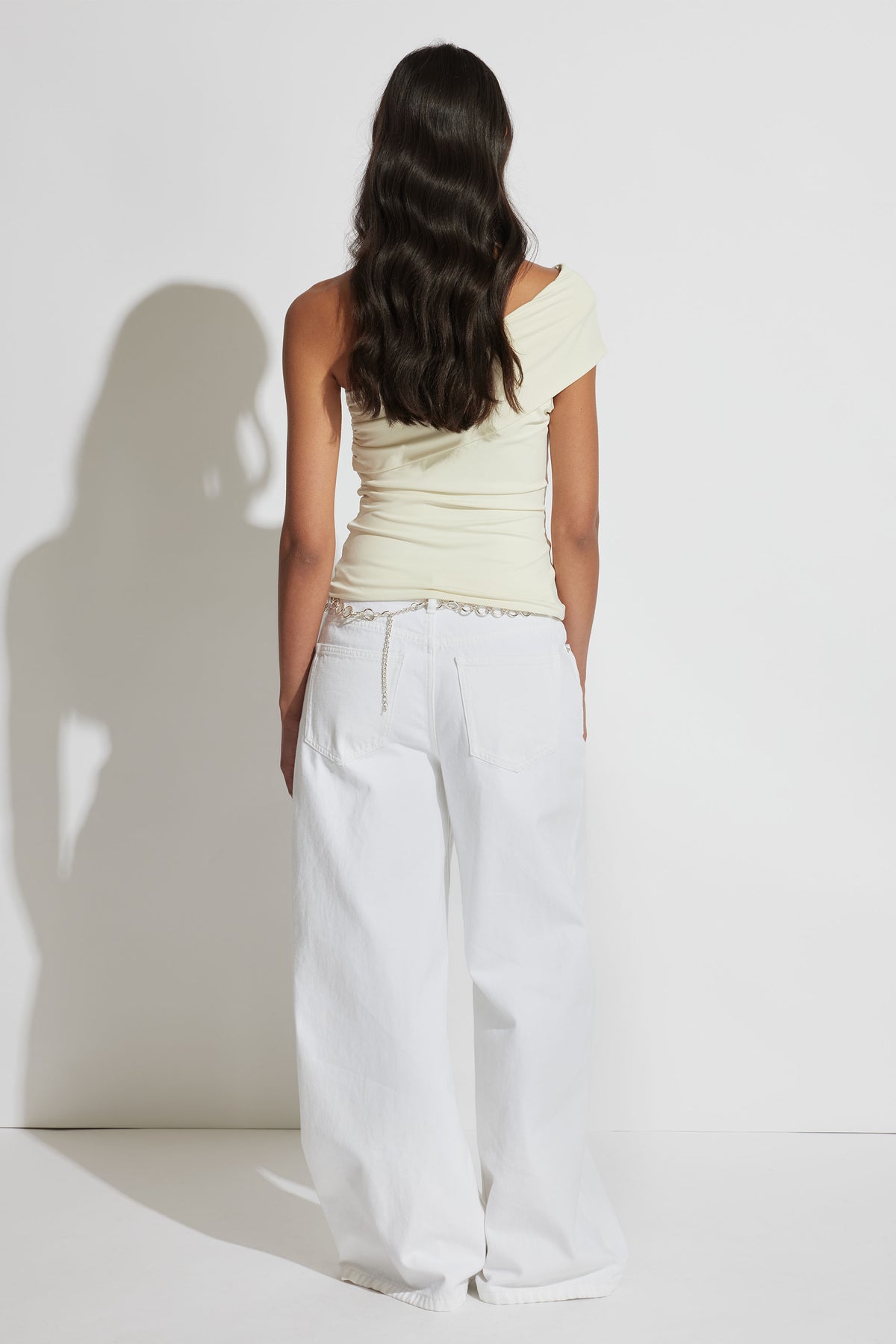 Woman wearing an off-white twisted one-shoulder top paired with relaxed wide-leg white denim jeans, seen from behind.