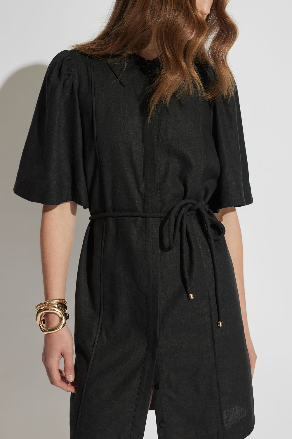 Woman wearing a black mini shirt dress with puff sleeves and a waist tie, accessorized with chunky gold bracelets.