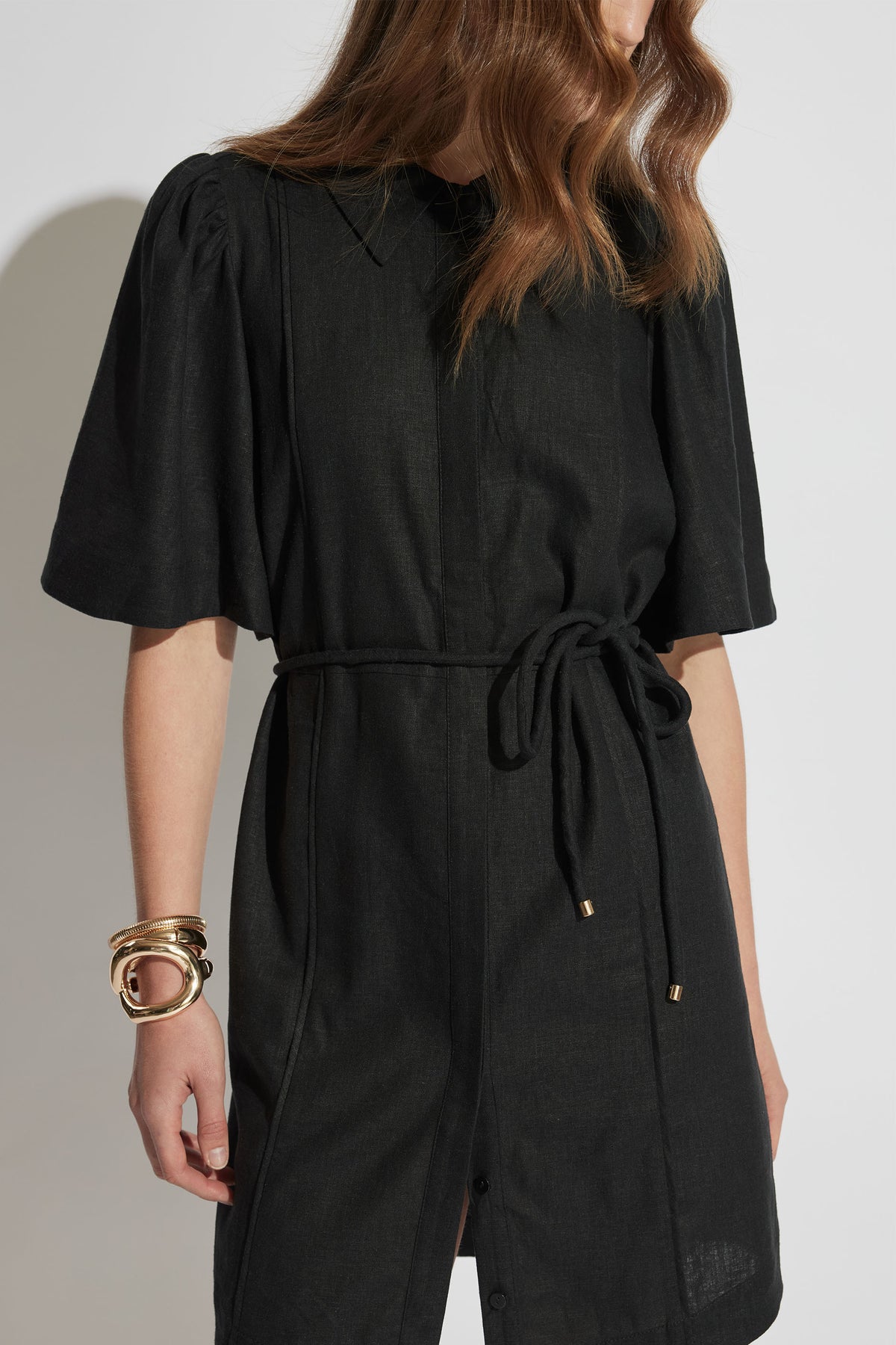 Woman wearing a black mini shirt dress with puff sleeves and a waist tie, accessorized with chunky gold bracelets.