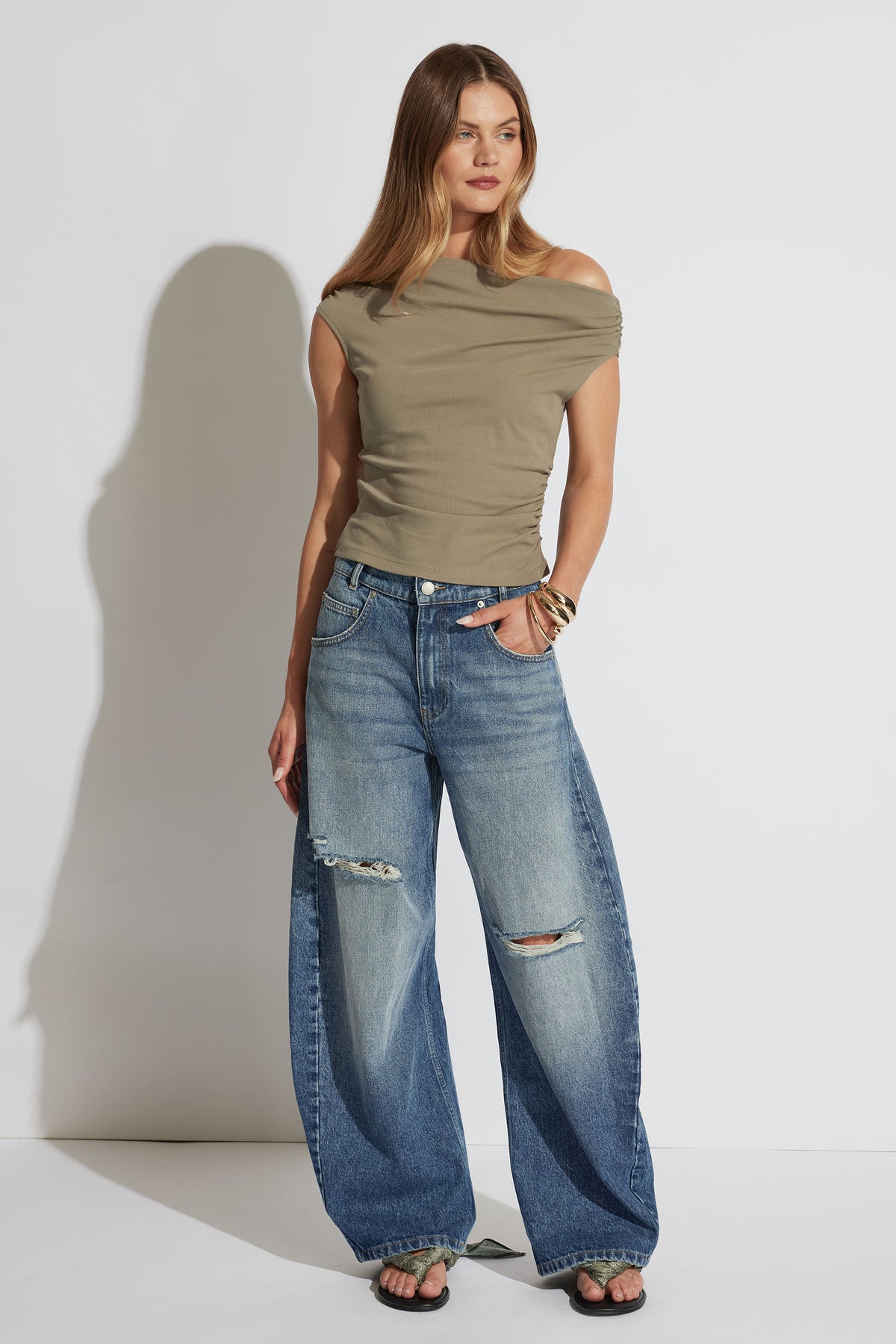 Woman wearing an olive off-shoulder fitted top paired with wide-leg distressed blue denim jeans and black sandals.