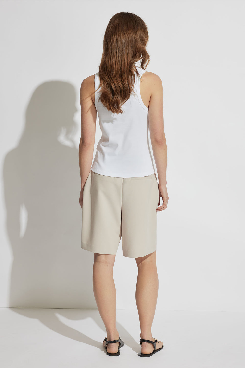 Woman wearing a white sleeveless tank paired with beige knee-length shorts and black sandals, shown from the back.