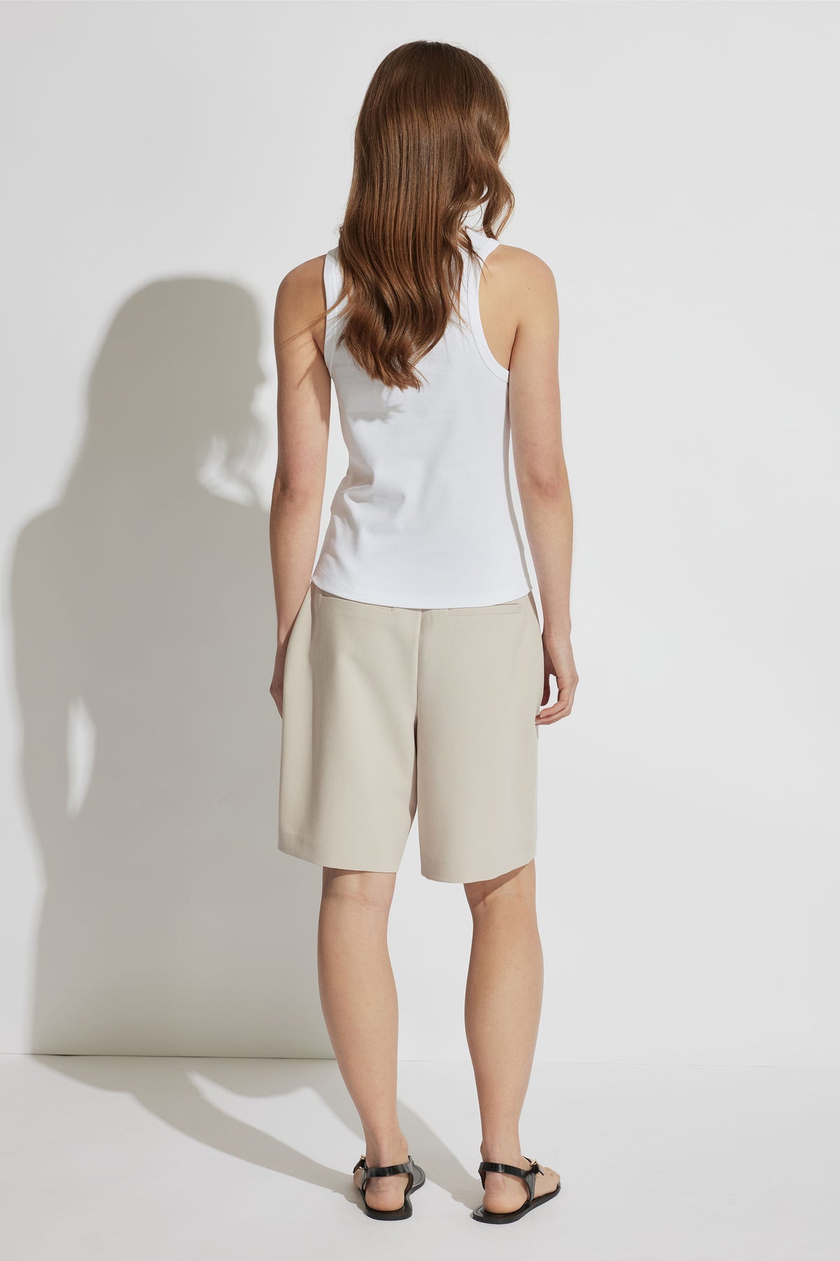 Woman wearing a white sleeveless tank paired with beige knee-length shorts and black sandals, shown from the back.