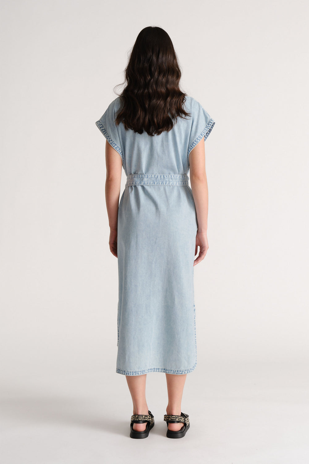 Woman wearing a light blue denim maxi dress with short sleeves and a belted waist, paired with black sandals.