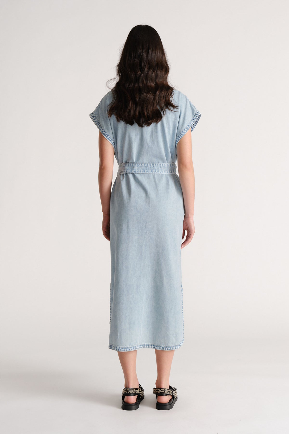 Woman wearing a light blue denim maxi dress with short sleeves and a belted waist, paired with black sandals.