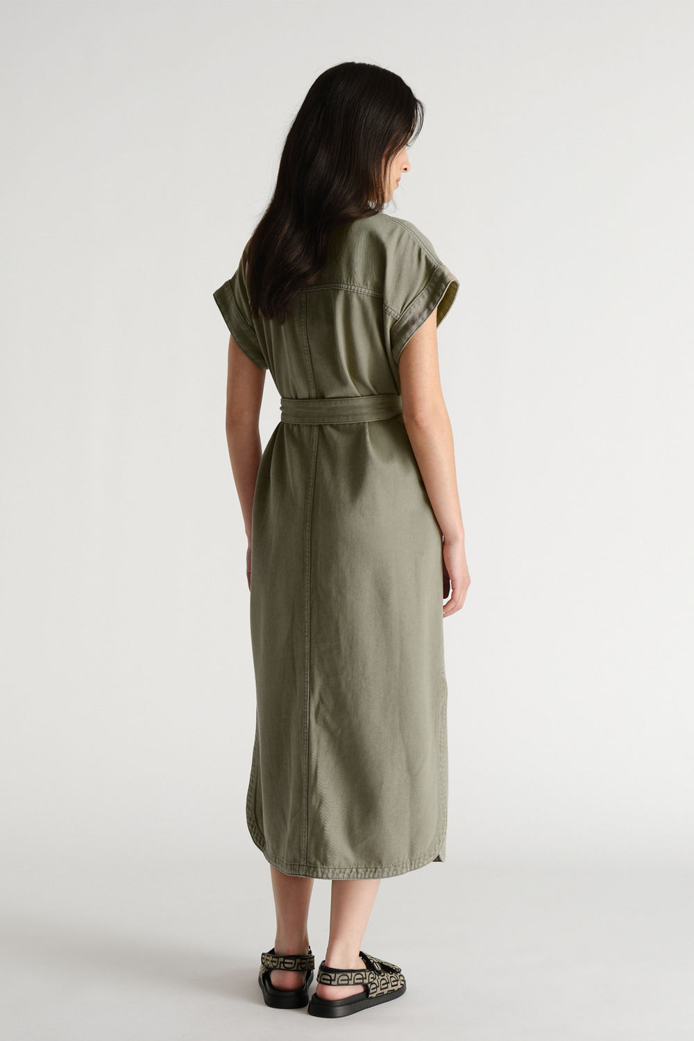 Woman wearing an olive green belted denim maxi dress with cuffed short sleeves and black slip-on sandals viewed from behind.