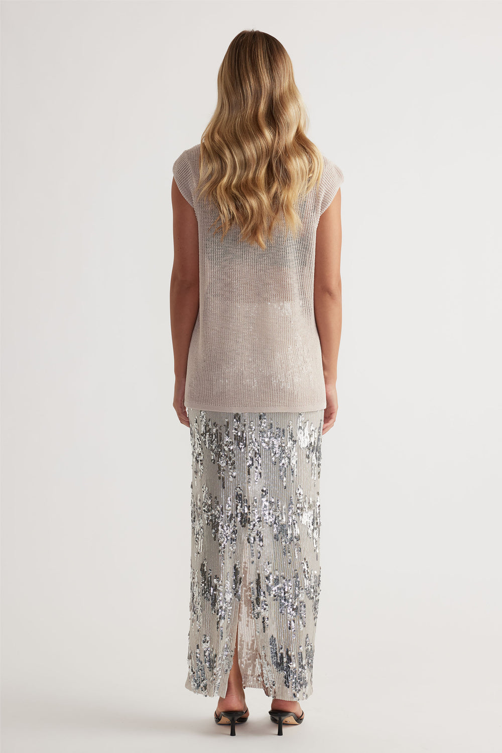 Woman with wavy blonde hair wearing a light neutral knit tank paired with a silver sequin maxi skirt and black heeled sandals