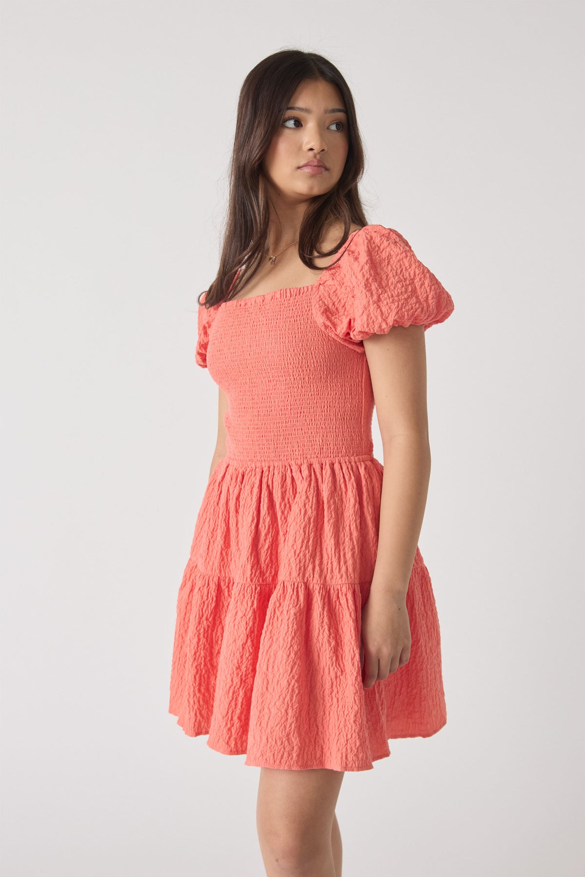 Young woman in a textured coral mini dress with puff sleeves and a smocked bodice, standing against a plain backdrop.