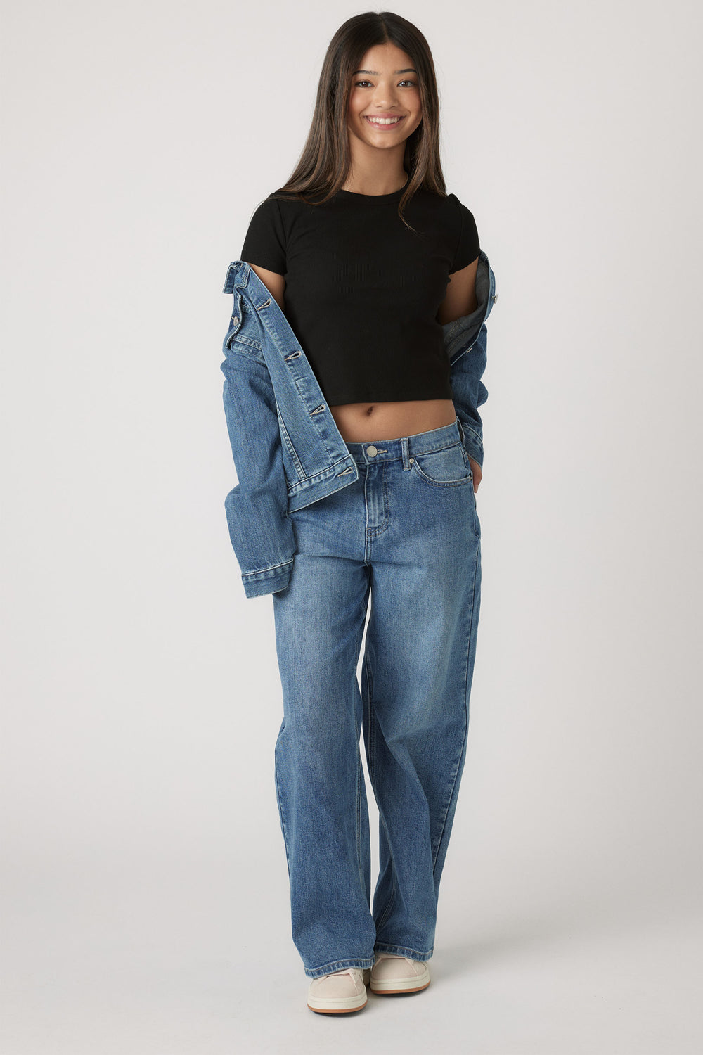 Teen girl wearing a black cropped tee and relaxed blue jeans, styled with a denim jacket casually draped off shoulders