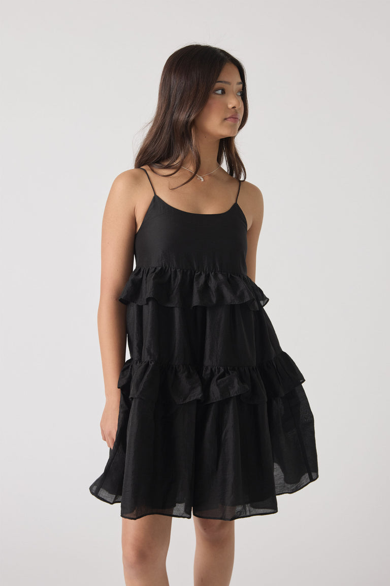 Girl wearing a black sleeveless dress with delicate ruffle tiers and thin spaghetti straps, looking to the side.