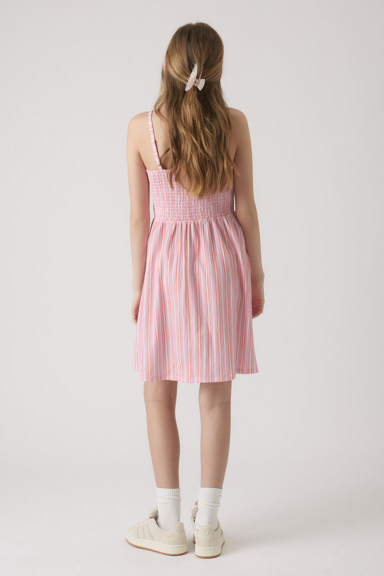 Girl wearing a pink striped shirred dress with spaghetti straps, paired with white socks, sneakers, and a clip in her hair.