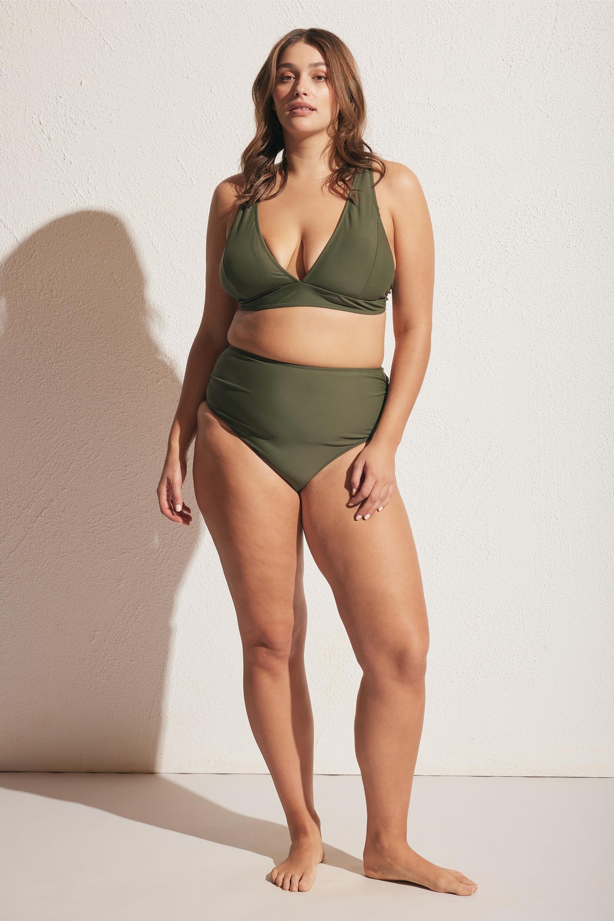 Woman standing barefoot wearing an olive green matte triangle bikini top and matching high-waisted bottoms against a white wall