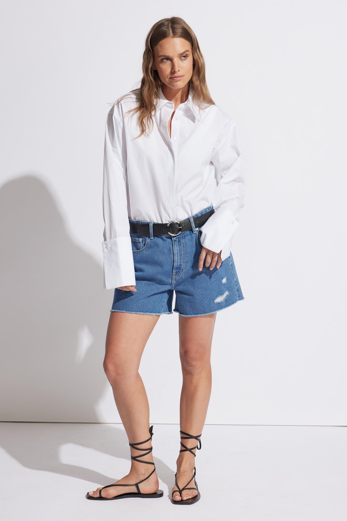 Woman wearing mid-blue distressed denim shorts with a black belt, paired with an oversized white shirt and black lace-up sandals
