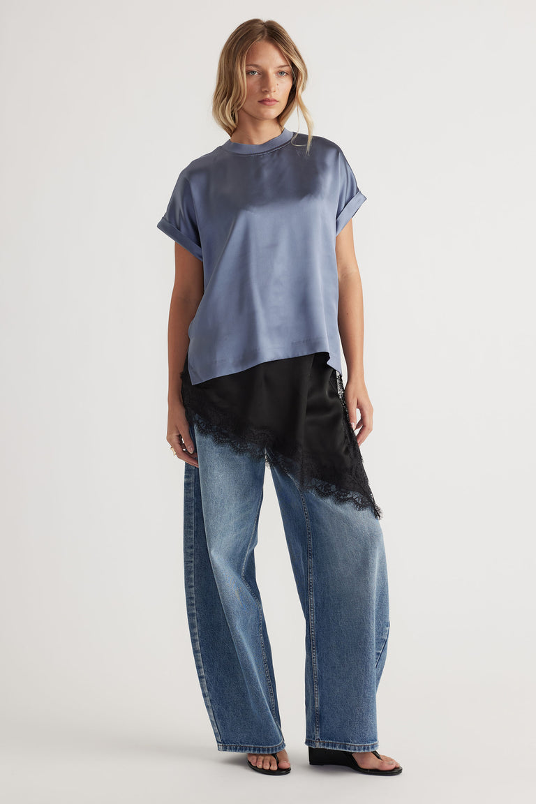 Woman wearing a loose steel blue satin tee with rolled sleeves, paired with wide-leg denim and lace-trimmed black layering piece