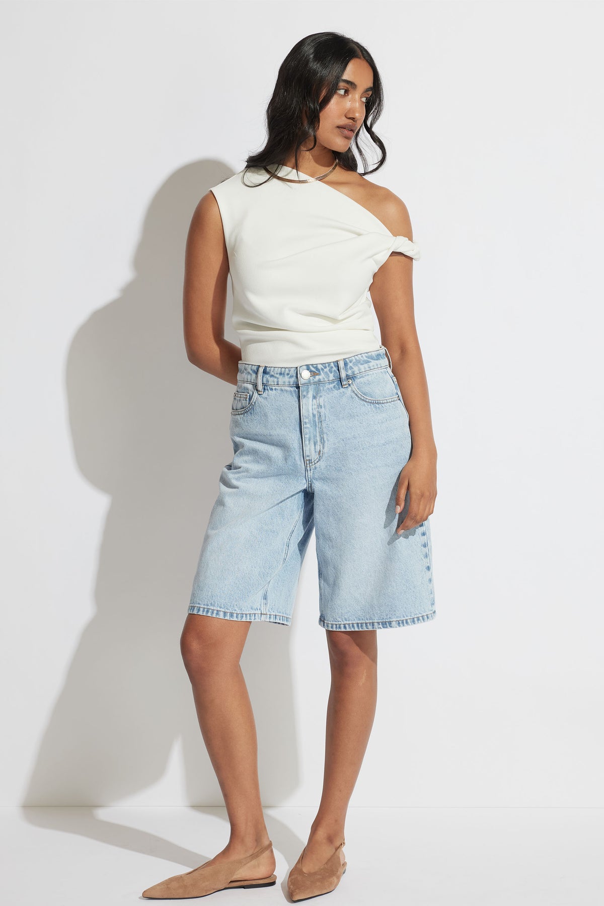 Woman wearing a white twist shoulder top with light wash knee length denim shorts