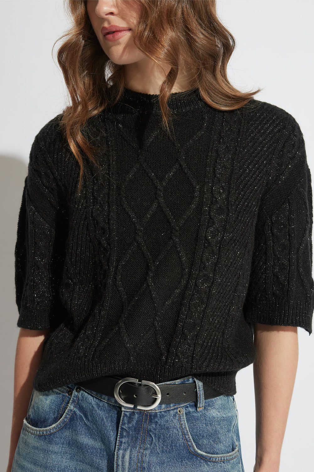 Woman wearing a textured black knit top with elbow-length sleeves, styled with a leather belt and casual blue jeans