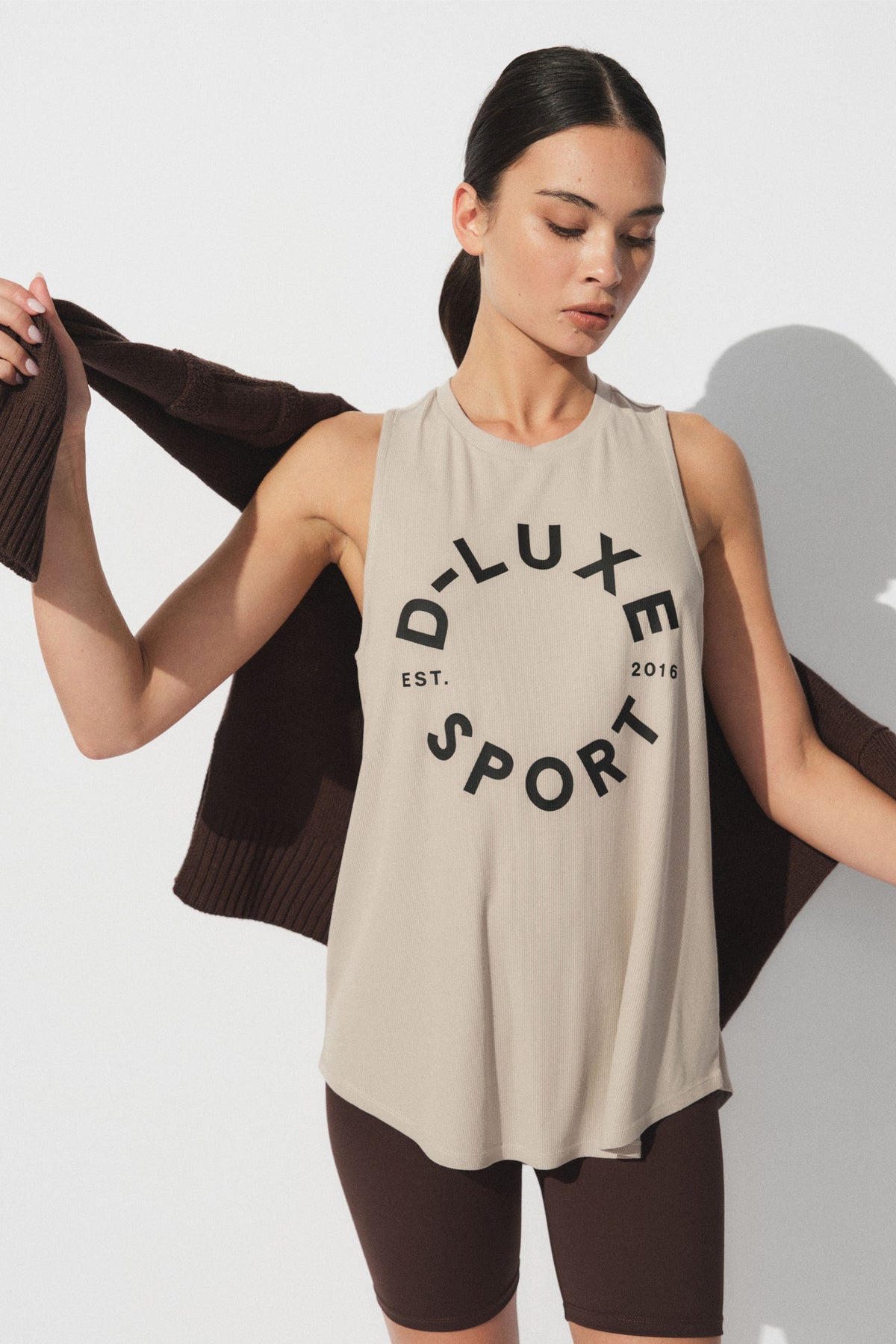 Woman wearing a sleeveless beige tank with bold graphic print paired with dark brown bike shorts and a knit sweater draped on shoulders