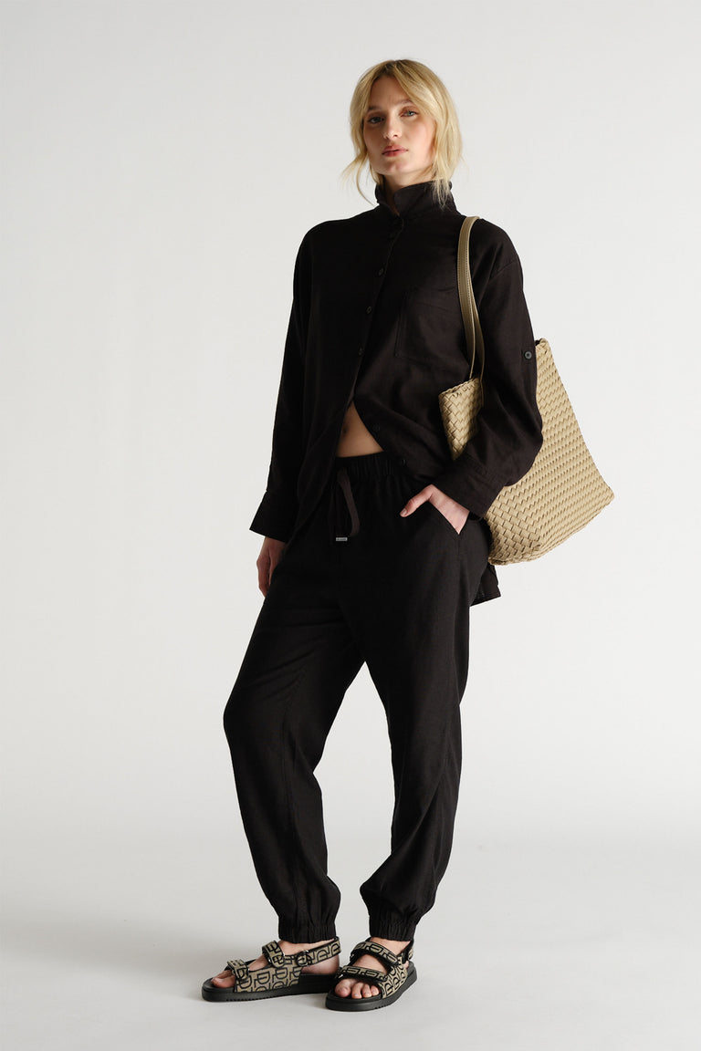 Woman wearing relaxed black linen blend joggers paired with a loose black button-up shirt and woven beige tote bag.