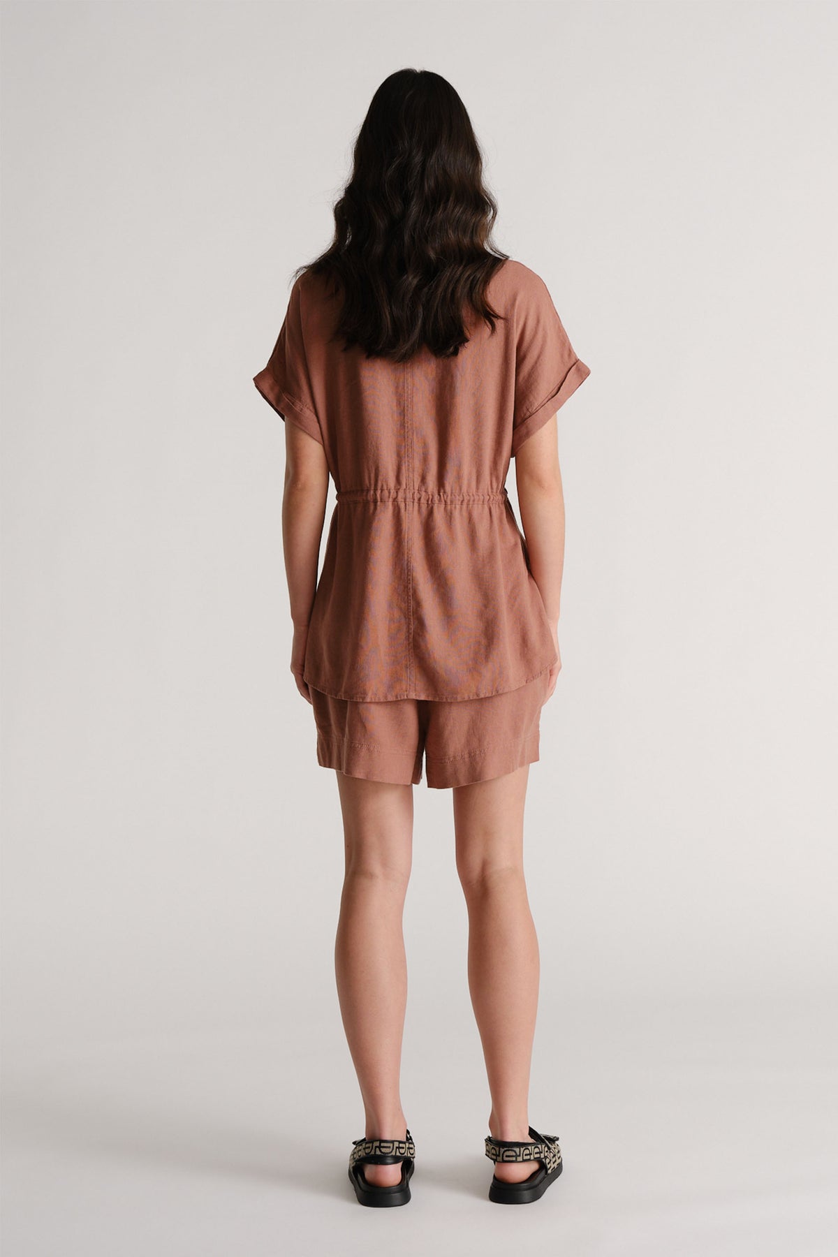 Woman wearing relaxed rosewood linen blend shorts with a matching loose top and patterned flat sandals viewed from behind
