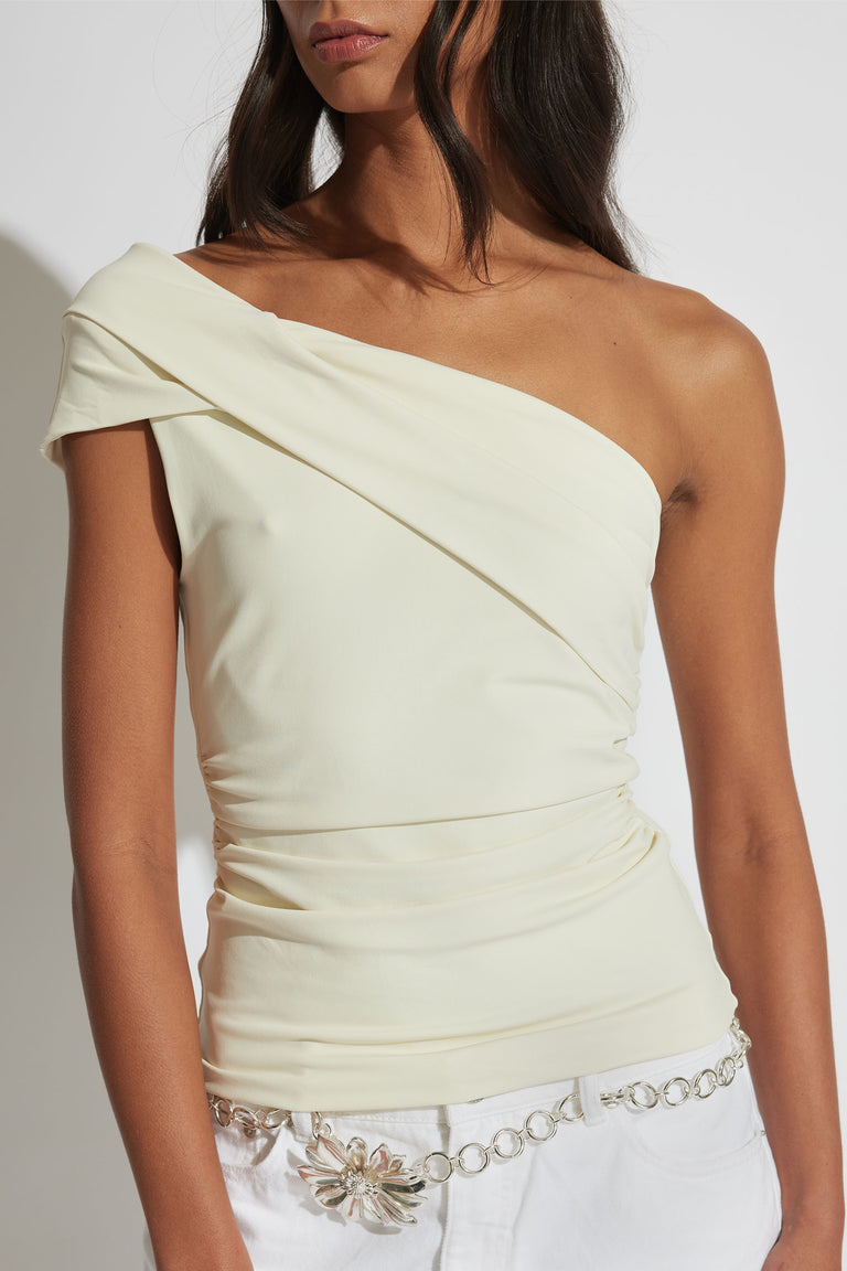 Woman wearing an ivory one-shoulder twist top paired with white pants and a silver floral chain belt.