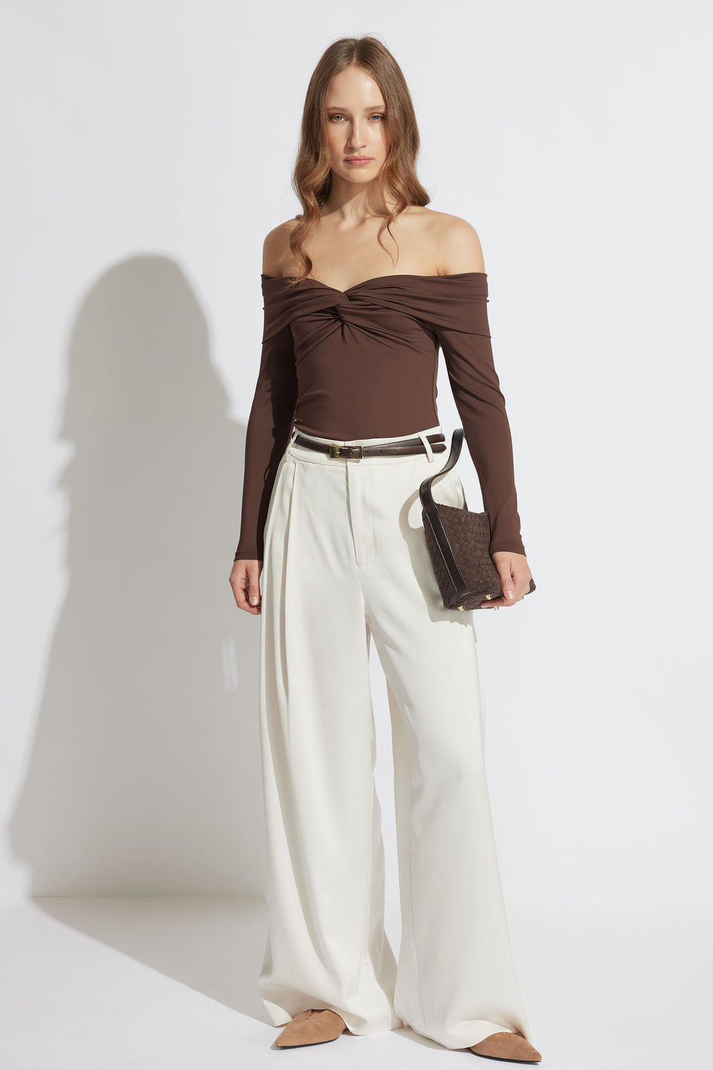 Woman in an off-shoulder brown twist-front long sleeve top paired with wide-leg cream trousers and a small handbag.
