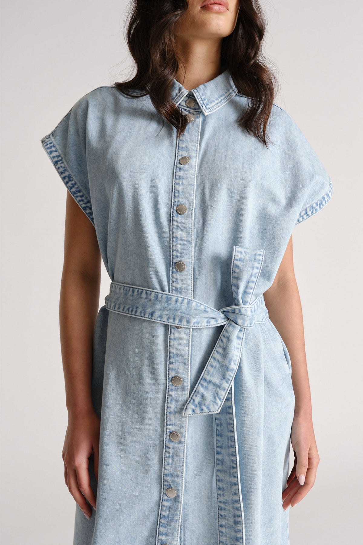 Woman wearing a light blue denim dress with capped sleeves, front buttons, and a matching fabric belt tied at the waist.