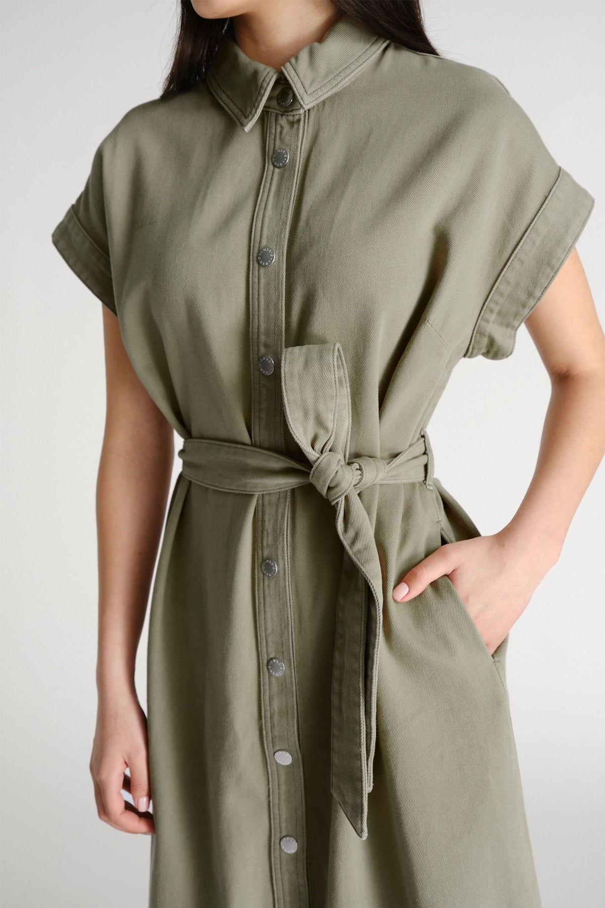 Woman wearing a thyme green belted shirt dress with rolled sleeves and silver snap buttons, hand casually in pocket.