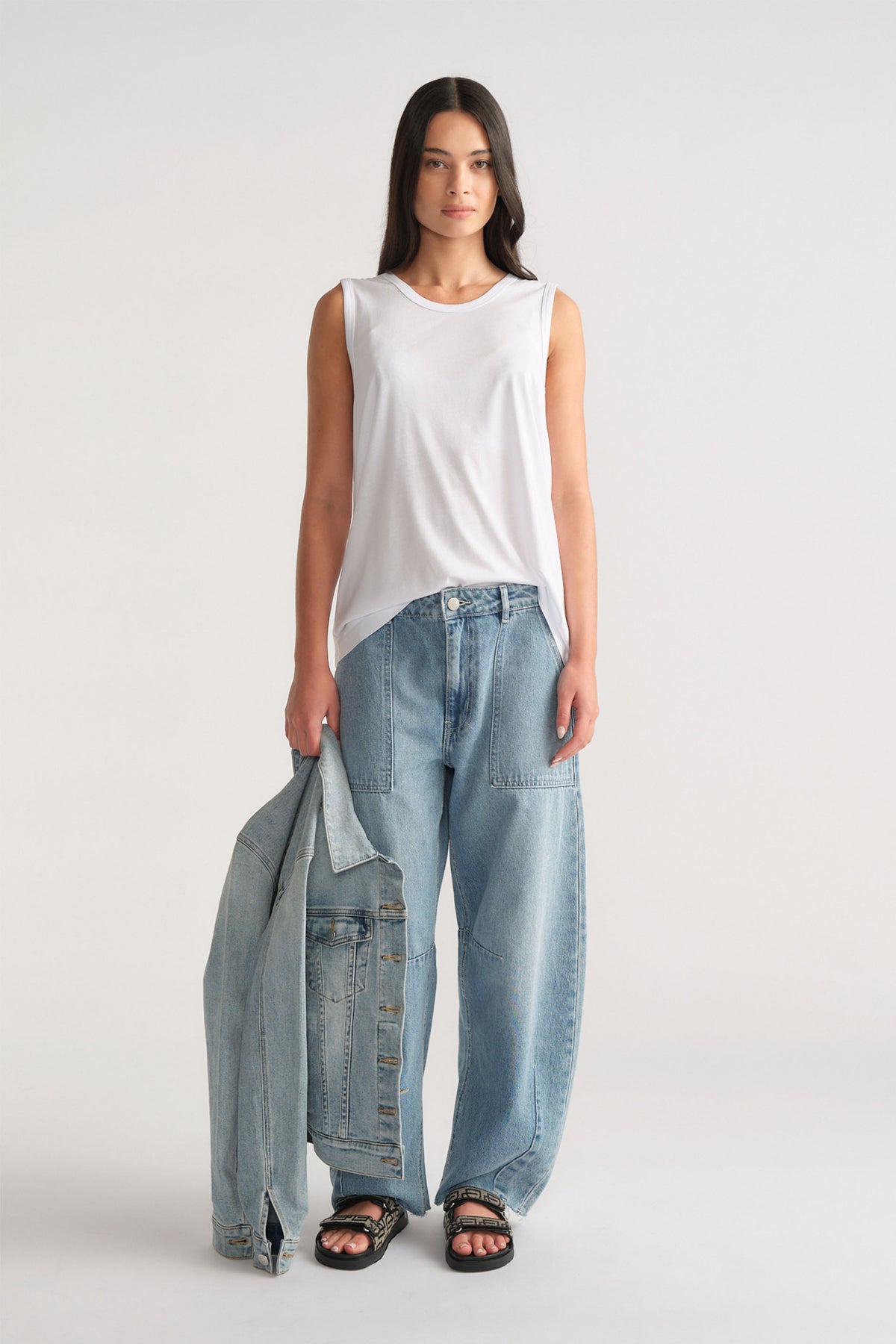Woman wearing a relaxed white sleeveless tank paired with loose light blue denim jeans and black sandals, holding a denim jacket.
