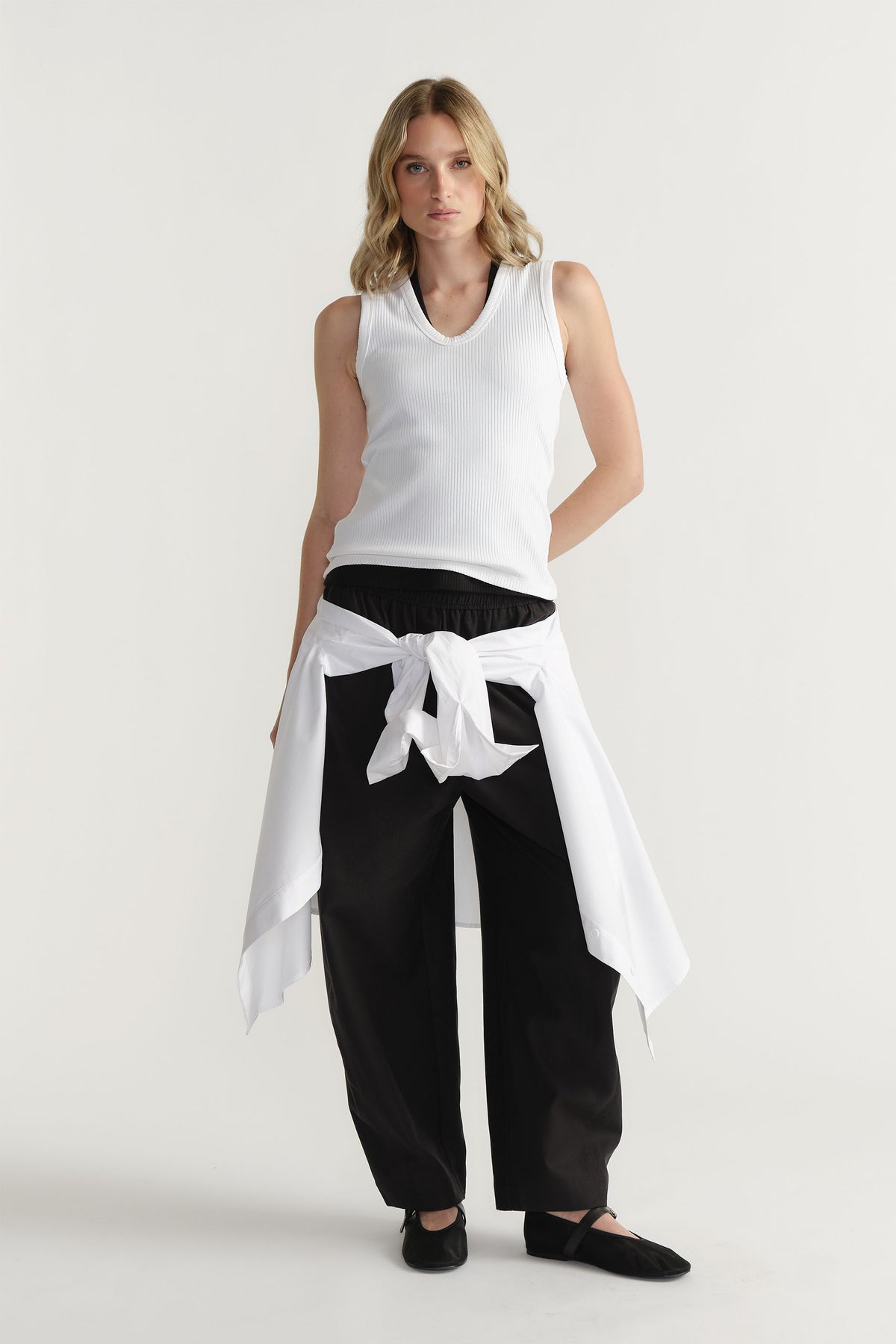 Woman wearing a white ribbed scoop tank layered over a black top, styled with black wide-leg pants and a tied white shirt waist.