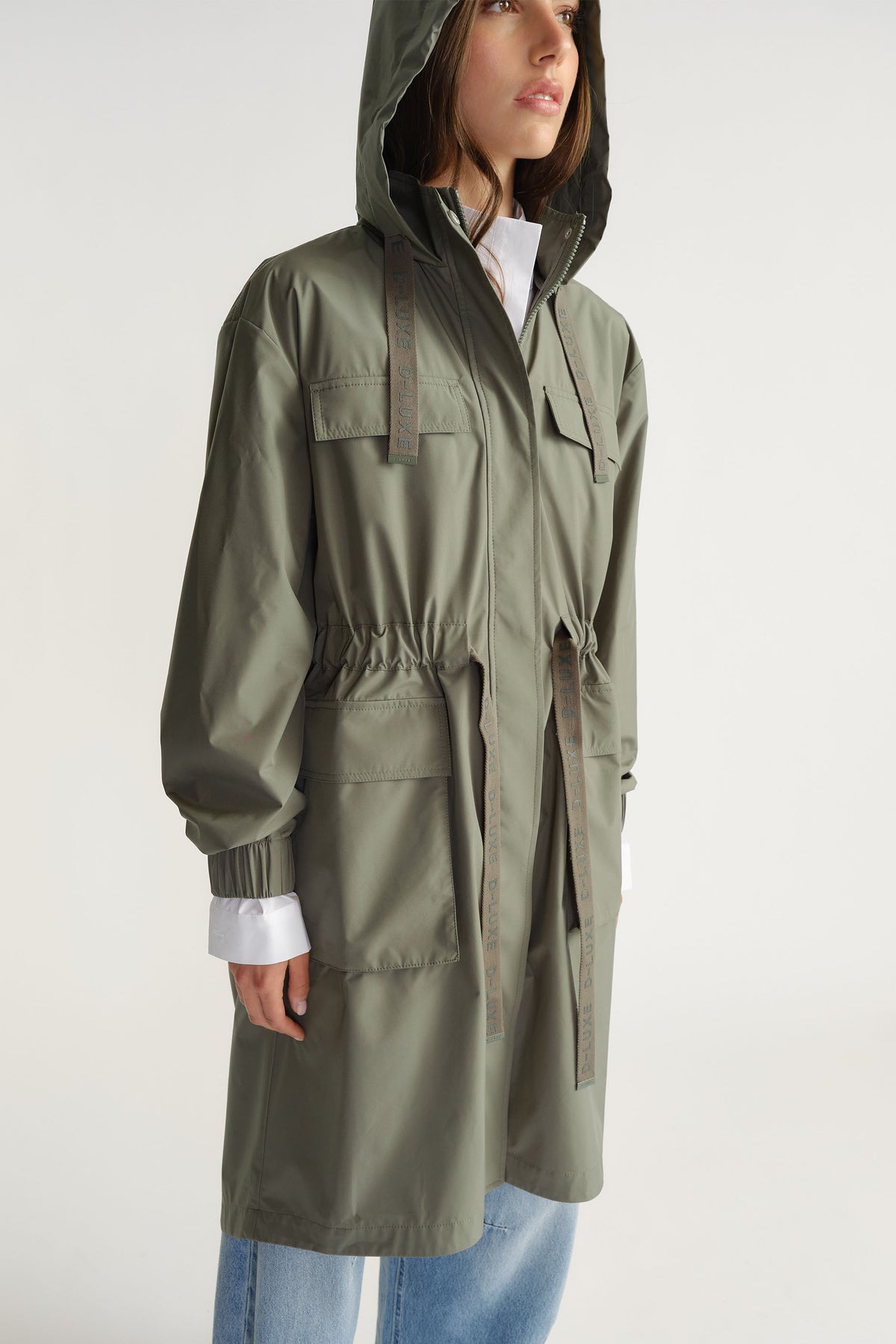 Woman wearing olive D-Luxe Rain Jacket with hood and adjustable waist paired with light denim jeans by Decjuba Australian brand