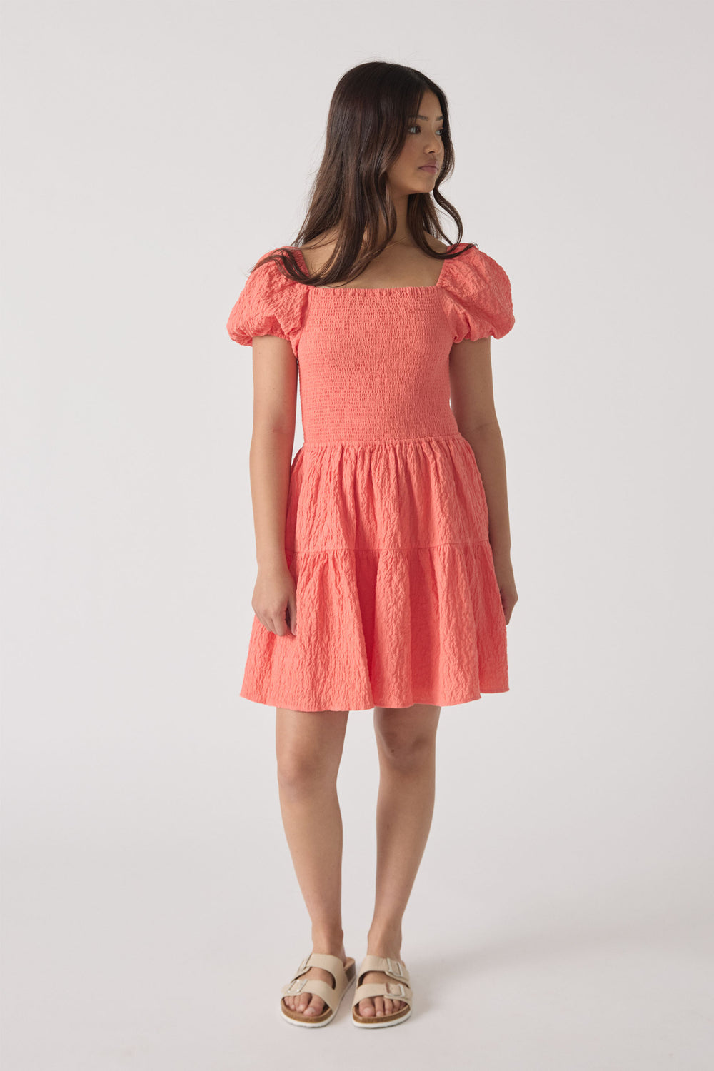 Girl wearing a coral pink mini dress with puff sleeves and textured tiered skirt, paired with cream sandals.
