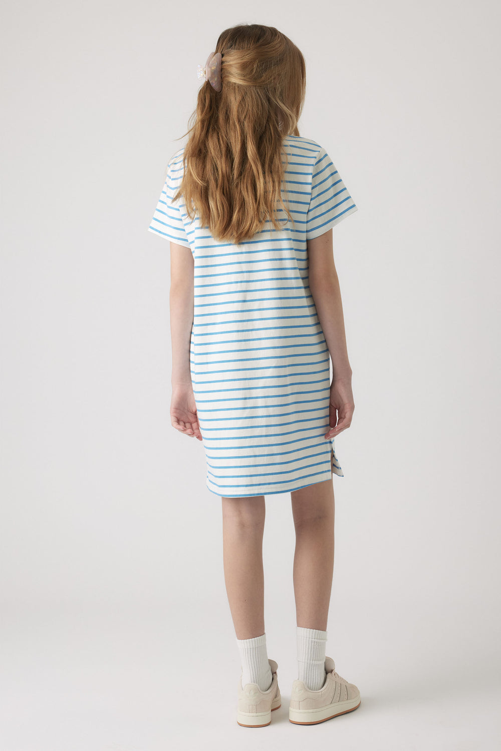 Young girl wearing a casual blue and white striped rugby dress with short sleeves and sneakers, facing away.