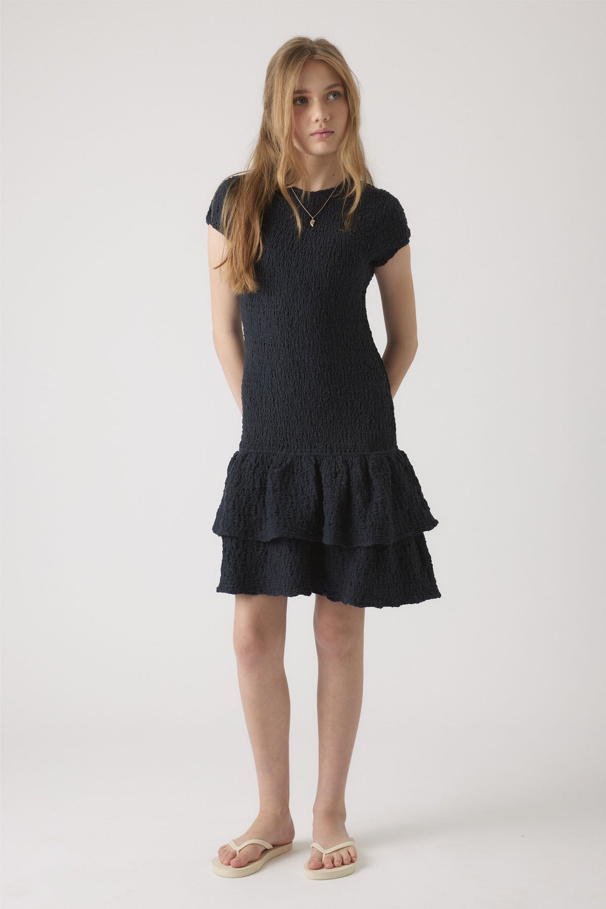 Girl wearing a navy shirred dress with cap sleeves and layered ruffle hem, styled with simple sandals in a neutral setting.