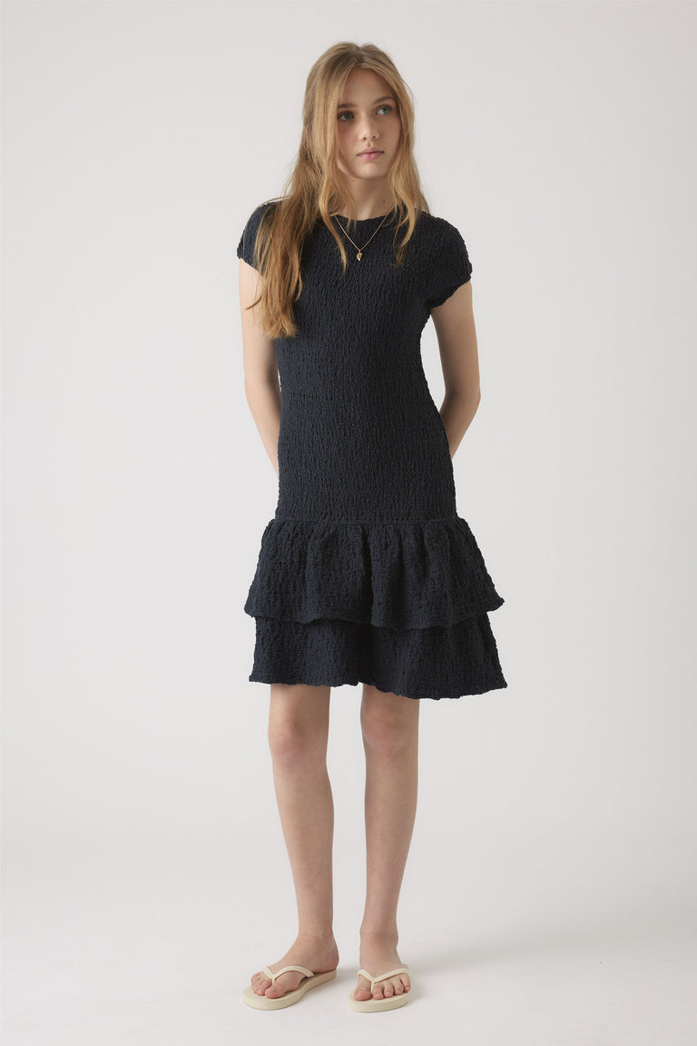 Girl wearing a navy shirred dress with cap sleeves and layered ruffle hem, styled with simple sandals in a neutral setting.