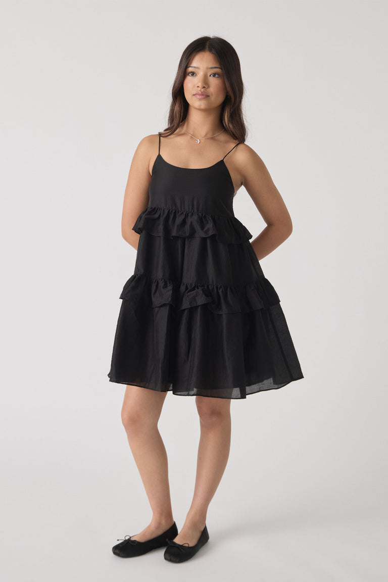 Girl wearing a sleeveless black ruffle dress with thin straps and ballet flats, standing with hands behind back.