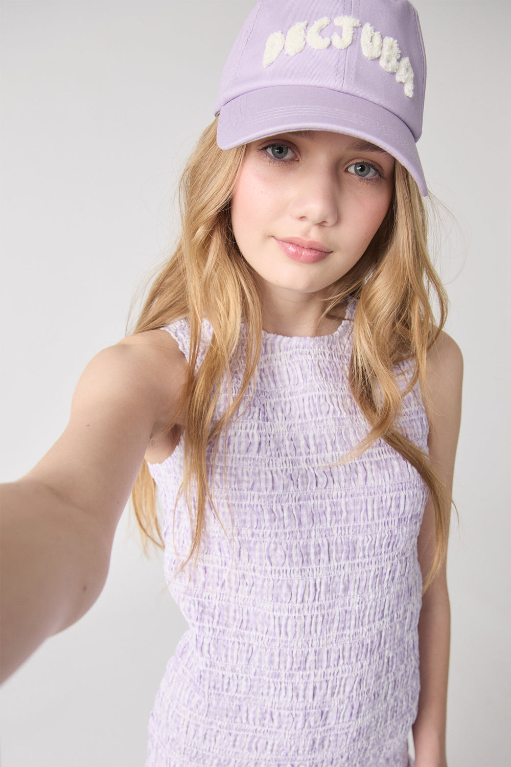 Girl wearing a lilac smocked sleeveless top and a matching purple cap with textured brand lettering.