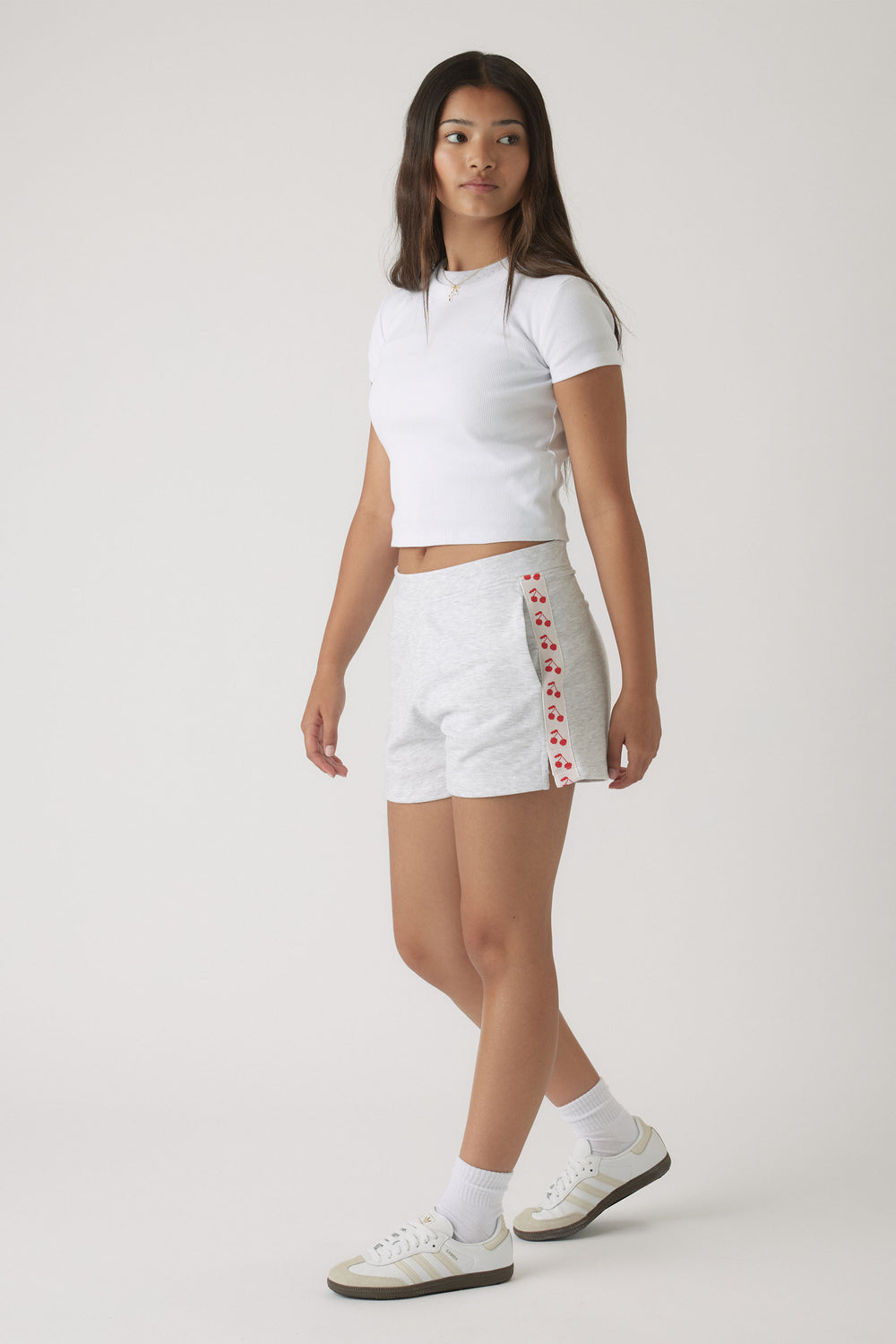 Teen girl wearing light grey sweat shorts with cherry print side stripes and a fitted white crop top with casual sneakers