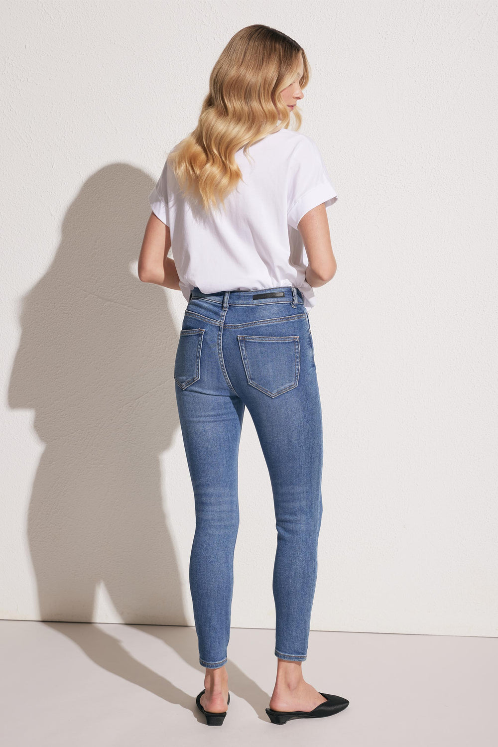 Woman with wavy blonde hair wearing mid-blue skinny jeans and a white short-sleeve tee, standing against a neutral wall