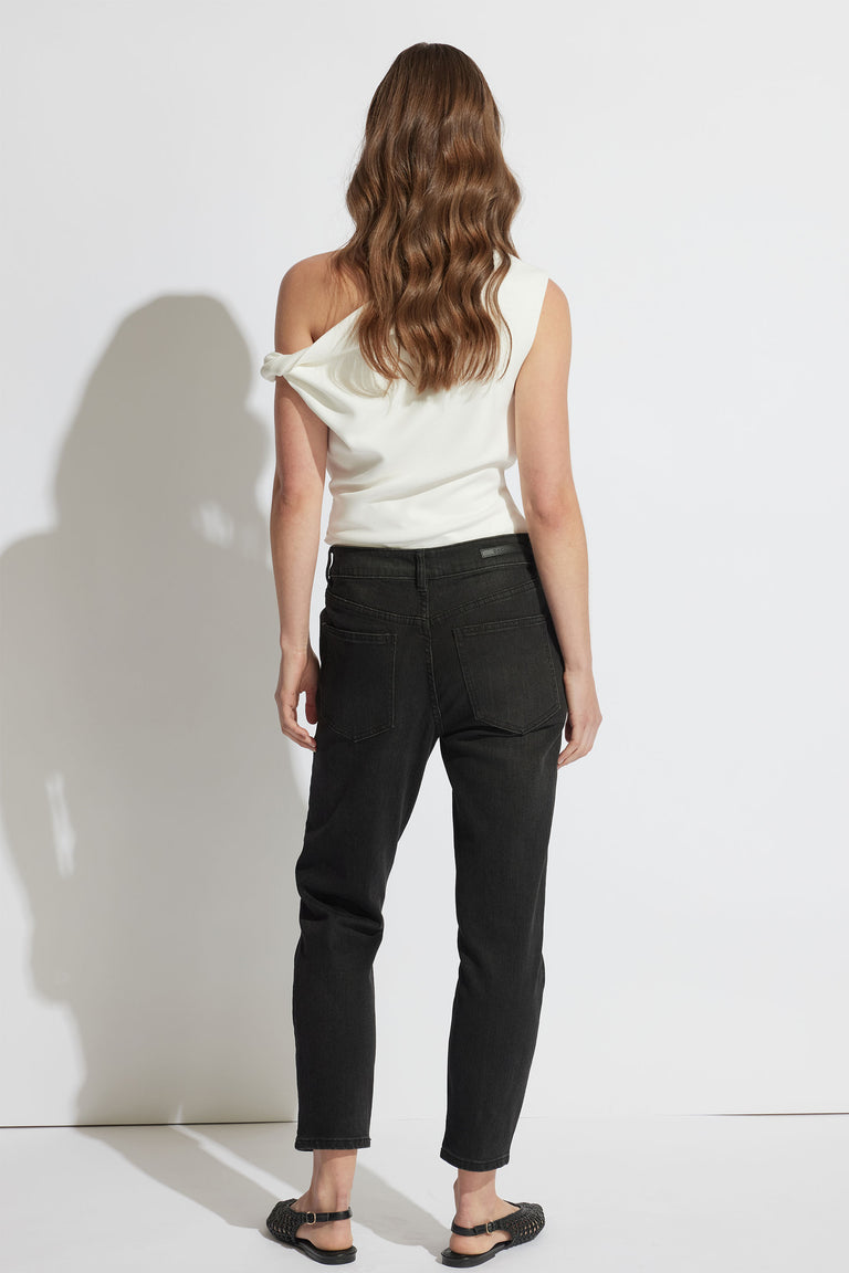 Woman wearing black relaxed fit jeans with a white off-shoulder top, standing against a white background.
